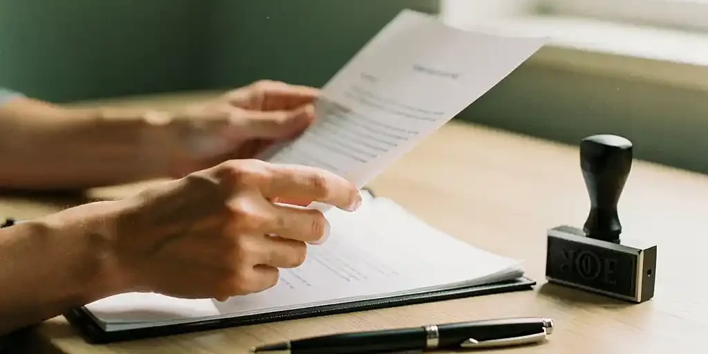 Handen van een ondernemer die een zakelijk contract doorleest op een houten bureau, met een bedrijfsstempel en pen ernaast.